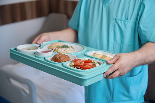 Cropped photo of male nurse with plastic meal tray in hands standing near hospital bed