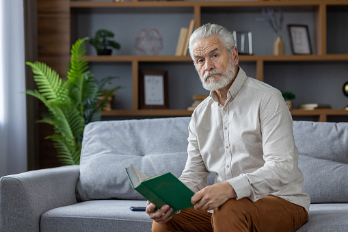 Portrait of a gray-haired senior man sitting at home on the couch, holding a book in his hands and looking seriously into the camera.