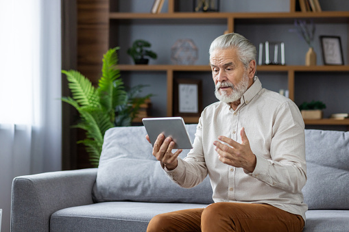 Senior gray-haired man communicates via video call from a tablet with a doctor, conducts an online consultation, explains the problem, sitting on the sofa at home.