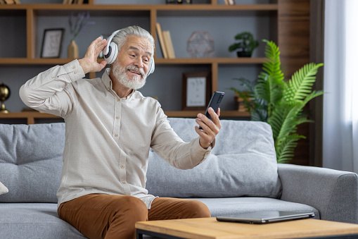 Senior man sitting on sofa at home and resting. Wearing white headphones, he listens to music from the phone and dances along.