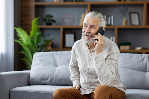 A gray-haired senior man is sitting on the sofa at home and talking on the mobile phone.