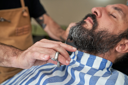 Anonymous barber using scissors for cutting hair of bearded male client while working in hairdressing salon