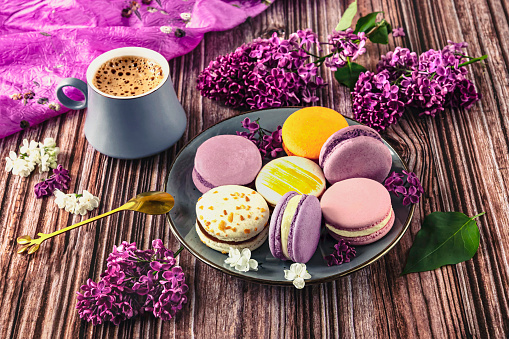 Colorful dessert macaroons on a plate with a spoon, gray cup of coffee on the table. Purple lilac as a decoration, top view