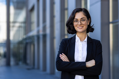 Young beautiful businesswoman in business suit smiling and looking at camera, portrait of successful woman outside office building, female worker in glasses with crossed arms.