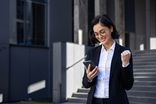 Young beautiful business woman in a business suit walking in the city with a phone in hands, a woman uses an application received a notification of a victory win, celebrates triumph and achievement.