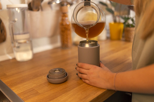 A woman pours hot herbal tea from a kettle into reusable cup in the kitchen while standing at the kitchen counter