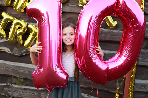 Cheerful little girl holds two big pink foil balloons, number 10 outdoors on wooden wall background with golden balloons inscription happy birthday. Ten years anniversary celebration party at home.