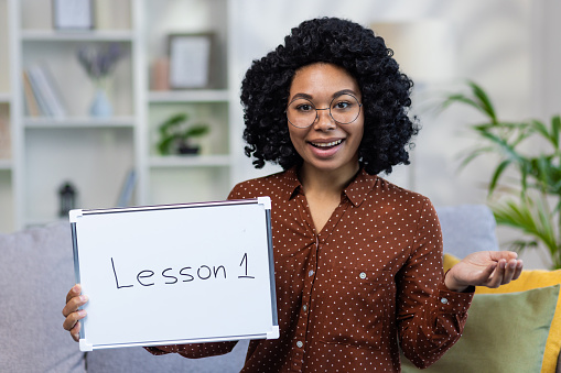 Portrait of a smiling African-American female teacher sitting at home on the sofa and teaching remotely, holding a blackboard with a lesson written in her hands. Close up photo.