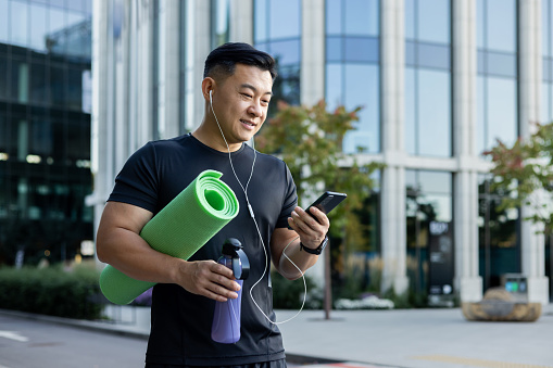 A young Asian sportsman man is standing on a city street in headphones, holding a phone, a yoga mat, and a bottle of water.