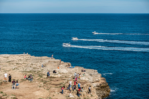 People enjoying the sunny weather on the rocky outcrop opposite the Cala Ponte beach in Polignano a Mare, Italy,
