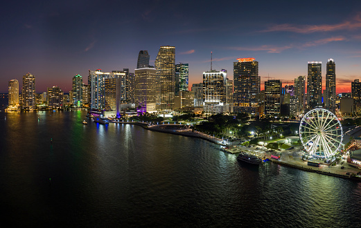 Aerial view of Skyviews Miami Observation Wheel at Bayside Marketplace with reflections in Biscayne Bay water and high illuminated skyscrapers of Brickell, city's financial center at night.