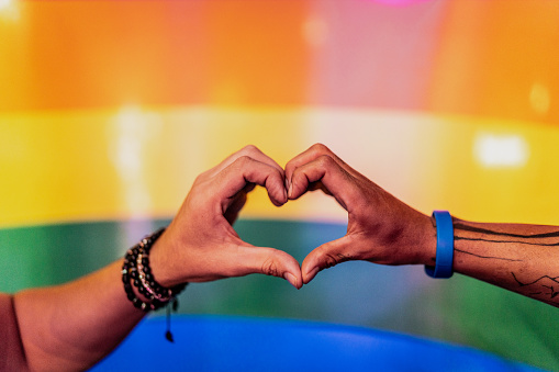 Close-up of a pride hands forming a heart shape with rainbow flag on background