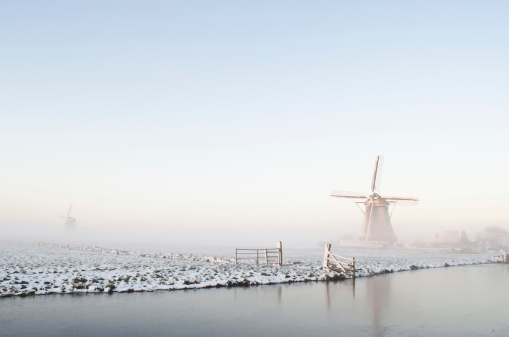 Several windmills in winter at the Kinderdijk, Holland. The water is frozen and all the land is covered with snow. There is a bridge over get water. The sun has just set.