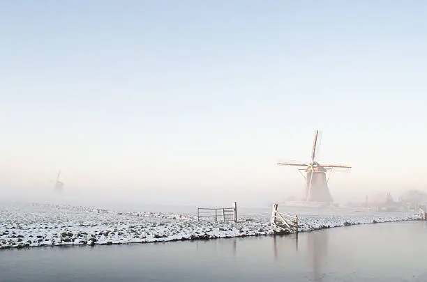 Typical winter landscape in Holland (the Netherlands) with a windmill, a canal, snow, ice and fog.