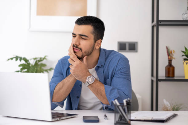 istock Close-up of young man feeling terrible toothache. Handsome young man touching his cheek while working on computer in home office, hurt and suffering from sensitive toothache. 1860506453