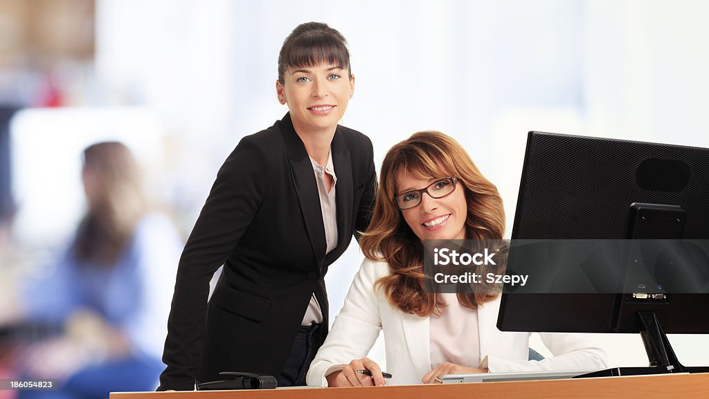 Businesswomen working in office Two smiling businesswomen working together in office with colleague in background Adult Stock Photo