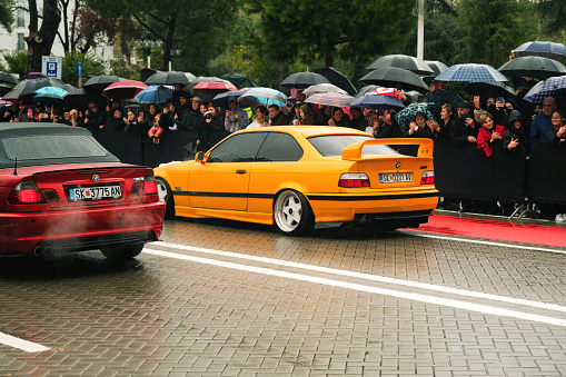 Tirana, Albania - November 28, 2023: On Avenue of Martyrs of the Nation, citizens watch a modified car parade on a rainy Independence Day, featuring a red and yellow BMW 3 E46 and labeled Cair Tuning Team