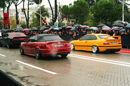 Tirana, Albania - November 28, 2023: On Avenue of Martyrs of the Nation, citizens watch a modified car parade on a rainy Independence Day, featuring a red and yellow BMW 3 E46 and a black colored dodge challenger labeled Cair Tuning Team