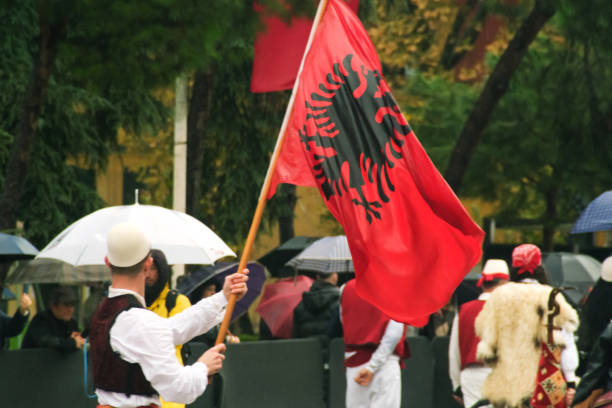 Tirana, Albania - November 28, 2023: On Avenue of Martyrs of the Nation, students and people in traditional attire enthusiastically march with Albanian flags on a rainy Independence Day, with spectators holding umbrellas and flags in the background.