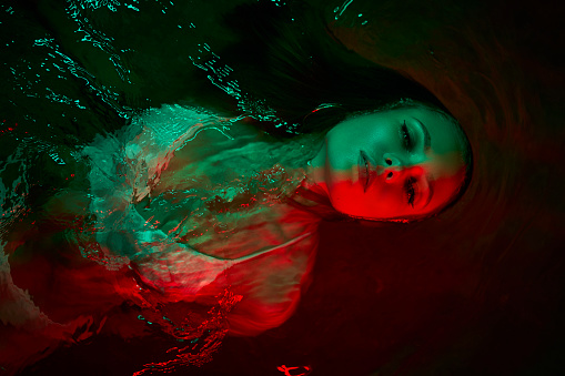 Woman floats in illuminated lagoon at night, submerged in water, green and red lights create ethereal ambiance. Female relaxed, fashionably dressed, tranquil nocturnal scene in serene body of water.