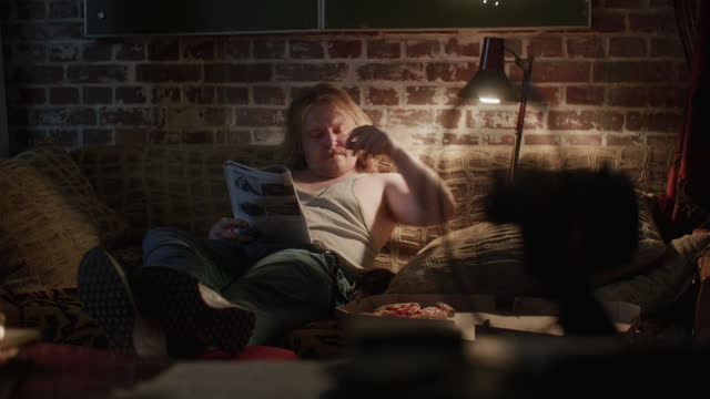 A lazy man sitting on a couch in his apartment reading newspapers