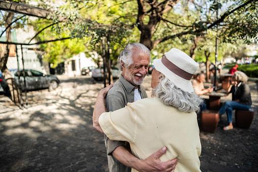 Elderly man looking at his elegant retired woman with love, warming her hands and enjoying her beauty, senior married couple dancing in autumn park, celebrating anniversary, affection