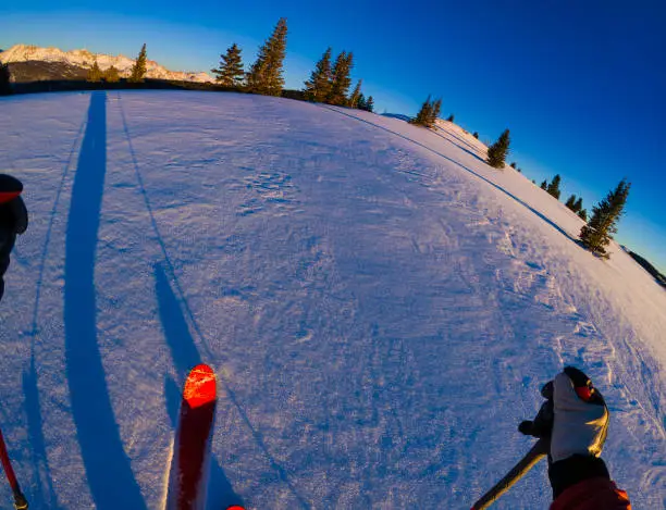 Backcountry Skier Skinning Mountain Ridge POV - Wide-angle, fisheye view from perspective of skier wearing red jacket and red skis. Ski touring up scenic ridge with mountain range bathed in warm alpenglow as backdrop. Outdoor adventure and recreation, personal fitness and healthy lifestyle.