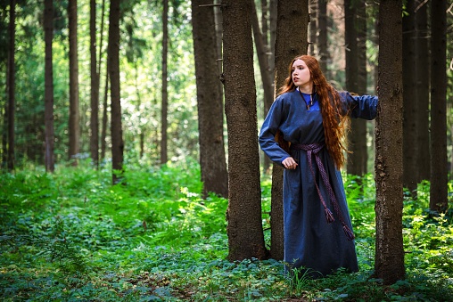 Red haired young woman in a linen blue dress is escape in the middle of the forest in the rays of the sun. The girl experiences emotions of despair left alone in the forest.
