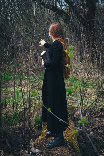 Gothic red haired woman in a black long dress in a gloomy forest. Mystical witch girl.