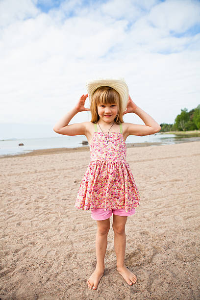 Smiling young barefoot girl wearing hat and pink patterned dress at beach