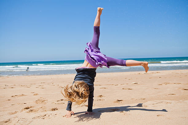 Cute young girl doing cartwheel in sand at beach with blue sky and ocean in background