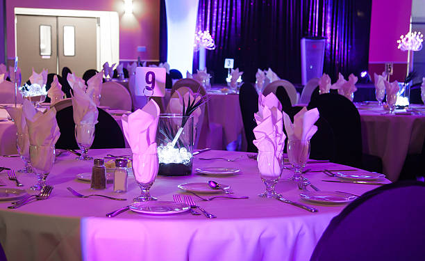 Elegant banquet setup with round tables, white chairs, floral centerpieces, and dishes in a hall with a stage prepared for an event.