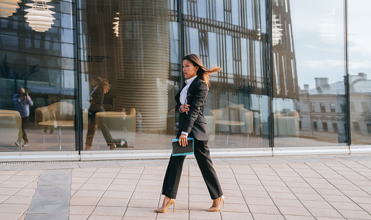 Focesed businesswoman in heels and a tailored suit strides past a reflective building, a picture of modern corporate determination and style.