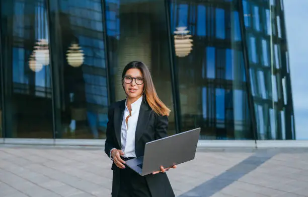 Ambitious businesswoman in eyeglasses and a sleek suit walks outside a modern glass building, laptop in hand, epitomizing professional grace and corporate success.