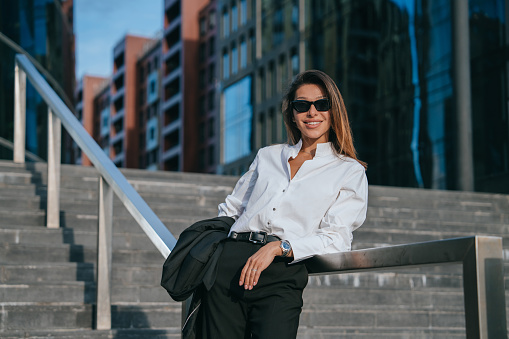 chic businesswoman in a smart suit poses with ease on city steps, her stylish sunglasses reflecting the dynamic architecture around her, showcasing a blend of elegance and corporate life.