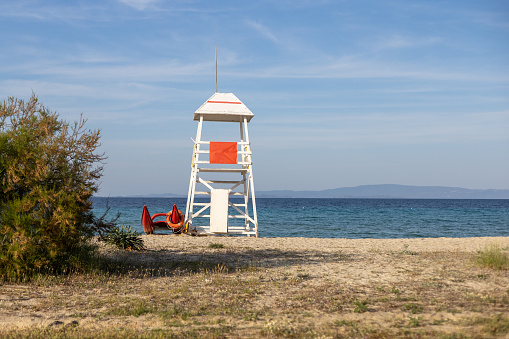 Empty white lifeguard chair on an empty beach with  orange life buoys hanging on the side.The chair stands against a clear blue sky in the sand.