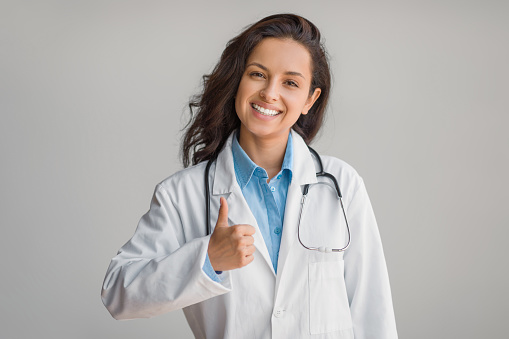 Happy female doctor in white coat, confidently giving thumbs up gesture, showcasing approval and positivity with a bright smile over light background