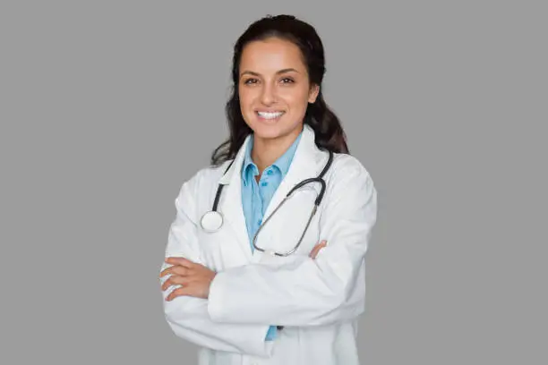 Confident female doctor in white coat and blue shirt, standing with arms crossed and stethoscope around her neck, exuding professionalism and warmth, grey background