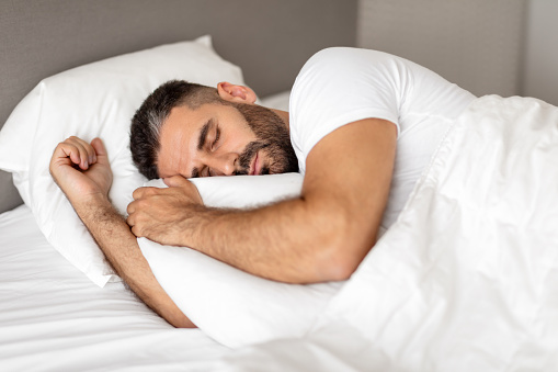 Man comfortably sleeping in bed wrapped in blanket, hugging white pillow in restful slumber and relaxation in modern bedroom setting. Shot of guy enjoying his nap and recreation