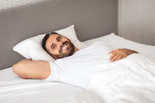 Relaxed European man happily lounging in comfortable bed, casually waking up in cozy modern bedroom, smiling as he lying with comfort under white blanket, enjoying good morning