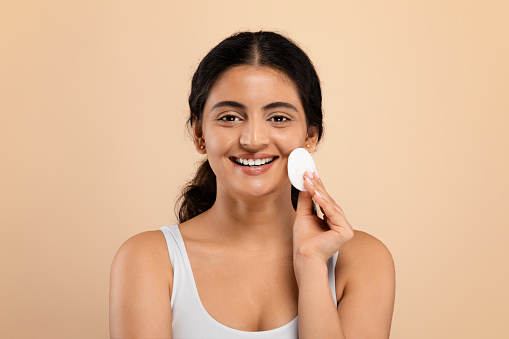 Skincare Routine. Smiling Attractive Indian Woman Cleansing Skin With Cotton Pad, Beautiful Eastern Lady Removing Makeup From Face, Standing Isolated On Beige Studio Background, Copy Space