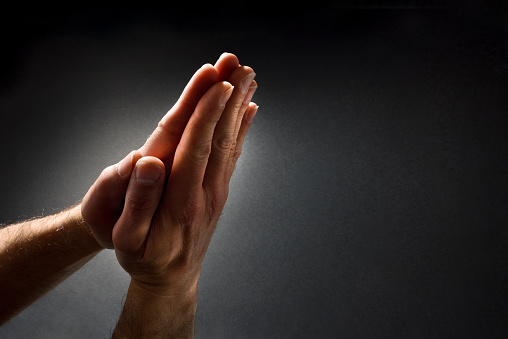 Praying man with hands with palms together contrasted with beam of light on dark isolated gradient background. Top elevated view.