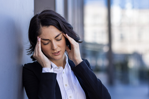 A close-up photo of a female office worker standing near her work on the street, leaning against the wall and holding her head from exhaustion.