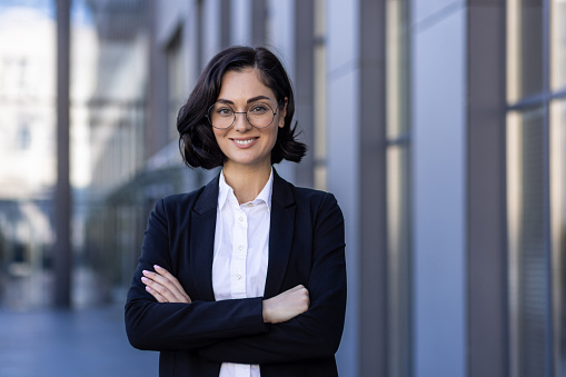Close-up portrait of a young businesswoman standing outside an office building with her arms crossed over her chest. Confidently and smilingly looking at the camera.