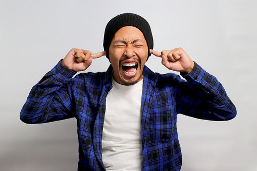 Angry young Asian man, in a beanie hat and casual outfit, is closing his eyes and covering his ears in response to noises. He appears to avoid hearing bad news, depicting stress under mental pressure