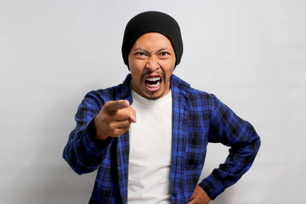 Angry young Asian man is yelling, pointing a finger, blaming all failures on someone Angry young Asian man in a beanie hat and casual shirt is yelling, pointing a finger, and expressing his frustration, blaming all failures on someone. Isolated on white background. asian suit man cross hand stock pictures, royalty-free photos & images