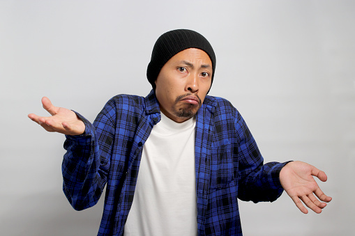 Confused young Asian man, dressed in a beanie hat and casual shirt is shrugging his shoulders in a gesture of cluelessness while standing against white background.