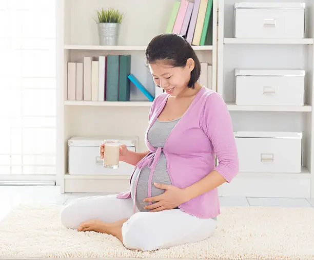 Asian 8 months pregnant woman enjoying a glass of soymilk as breakfast,  relaxing at home.