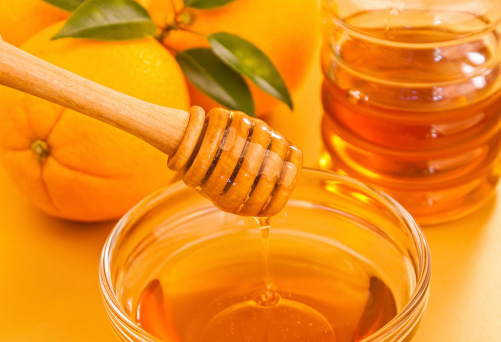 Bowl of orange honey with wooden dipper drizzler and a jar and oranges in the background