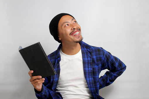 An enthusiastic young Asian student, dressed in a beanie hat and casual shirt, holds a book while looking at an empty space meant for copy or text, seemingly lost in imagination or contemplation